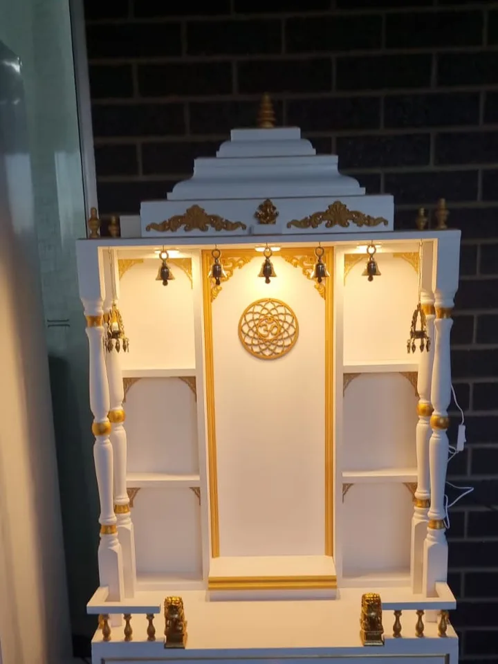 White and gold mandir lit from within, with gilded pillar bands and a carved om roundel