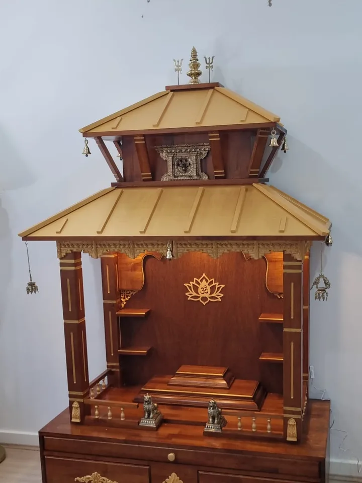 Two-tier teak mandir photographed at an angle, showing the carved eave fringe and lotus om panel